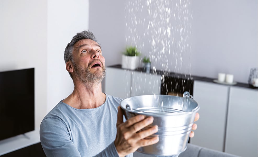 Man wearing gray shirt holding a bucket under a leaking ceiling