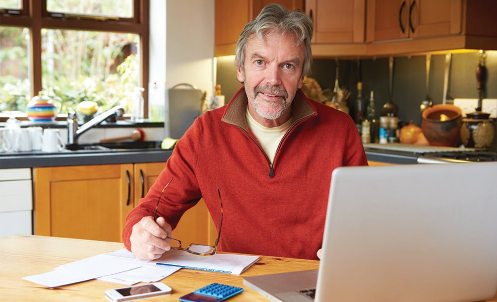 Smiling man wearing red sweater working on paperwork at laptop while sitting in kitchen