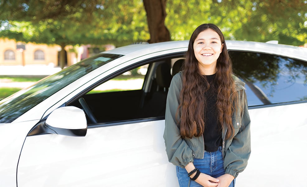 Teenage girl standing next to white car parked under trees