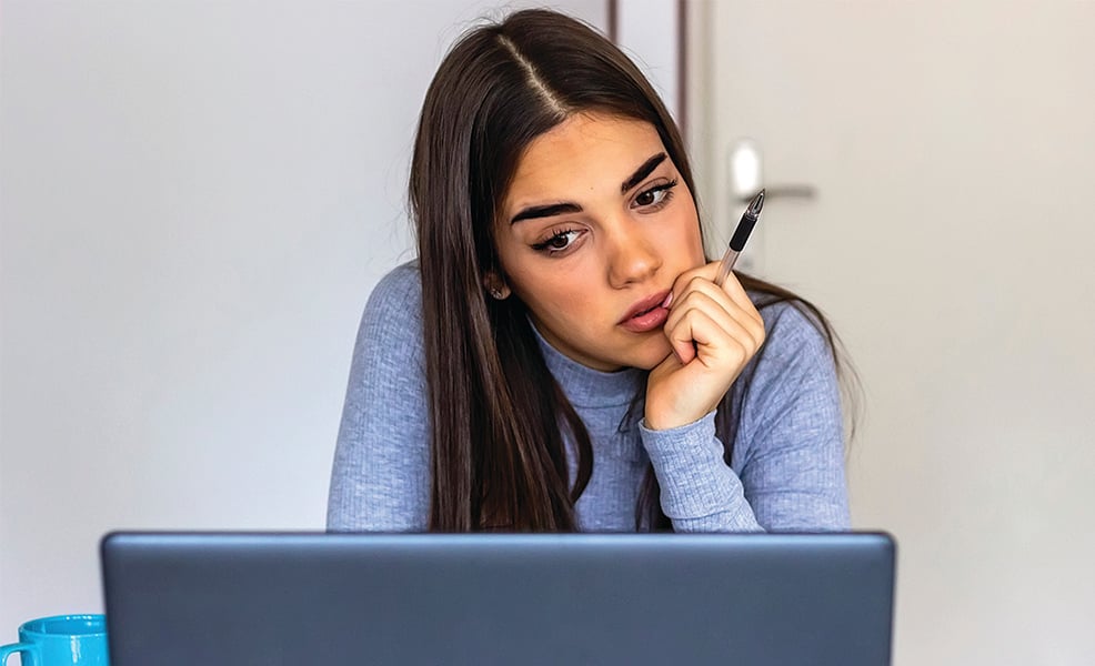 Woman with long hair wearing light blue sweater looking concerned at laptop 